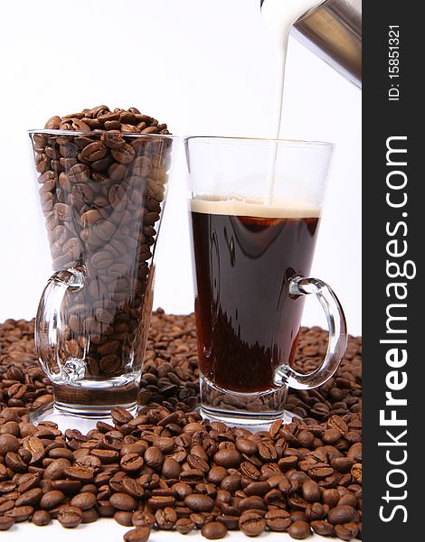 Making of caffe latte:glass of black coffee with milk being poured into it, with a glass of beans surrounded by coffee beans