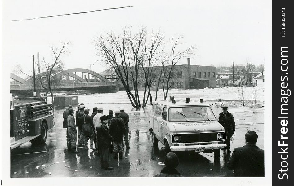 A photograph showing police, firefighters and city workers standing next to the Moira River during the 1981 flood in Belleville, Ontario. The Bell Shirt Company and the Lower Bridge can be seen in the background. This photo was taken by Elaine Preston. A photograph showing police, firefighters and city workers standing next to the Moira River during the 1981 flood in Belleville, Ontario. The Bell Shirt Company and the Lower Bridge can be seen in the background. This photo was taken by Elaine Preston.