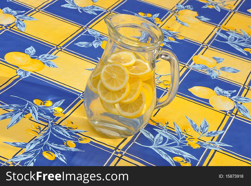 Crystal glass pitcher with lemon slices in water on yellow and blue plastic tablecloth. Crystal glass pitcher with lemon slices in water on yellow and blue plastic tablecloth