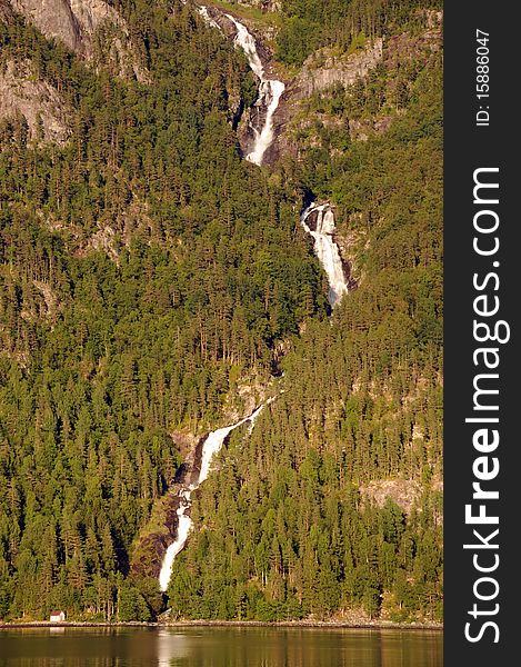 A waterfall tumbles through forested slopes down to a lonely house on Hardangerfjord, Norway. A waterfall tumbles through forested slopes down to a lonely house on Hardangerfjord, Norway