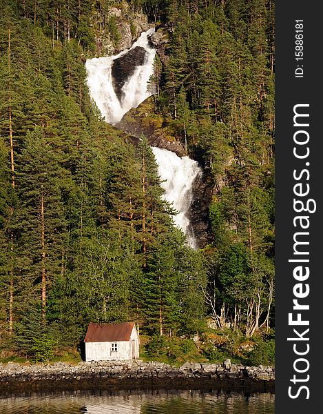 A waterfall tumbles through forested slopes down to a lonely house on Hardangerfjord, Norway. A waterfall tumbles through forested slopes down to a lonely house on Hardangerfjord, Norway