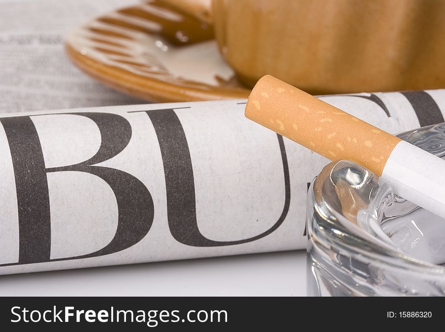 A filtered cigarette resting on a glass ashtray next to a brown cup and plate and a newspaper.