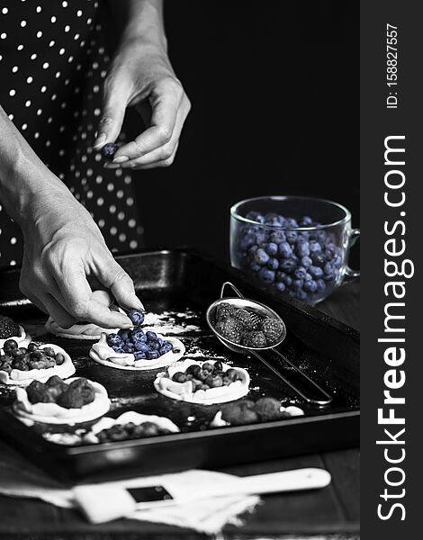 Chef`s hand is preparing cookies from puff pastry with raspberries and blueberries on a baking sheet on a dark background