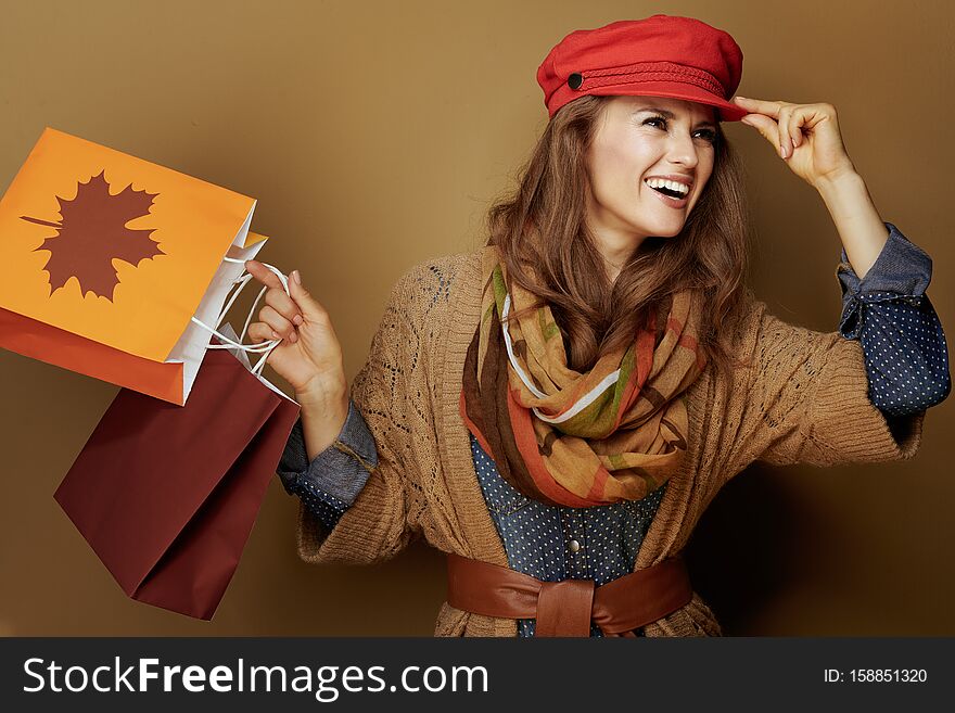 Smiling modern middle age woman in red cap, scarf, jeans shirt and cardigan with autumn shopping bags looking aside  on brown background. Smiling modern middle age woman in red cap, scarf, jeans shirt and cardigan with autumn shopping bags looking aside  on brown background