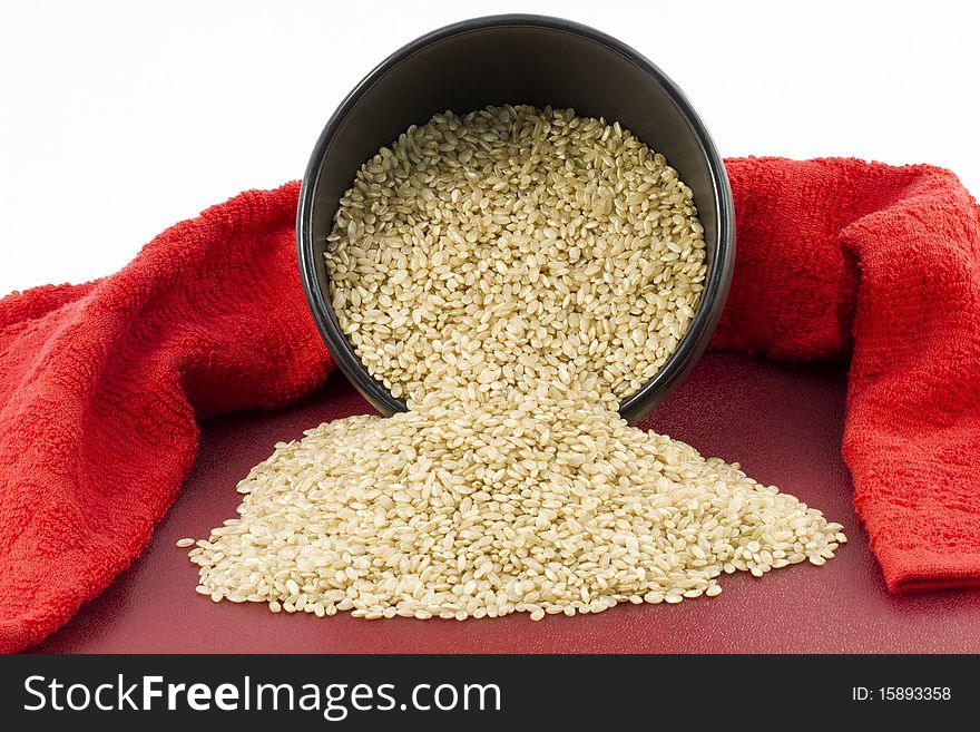 Brown rice spills from a dark ceramic bowl onto red kitchen board surrounded by red kitchen towel. Brown rice spills from a dark ceramic bowl onto red kitchen board surrounded by red kitchen towel