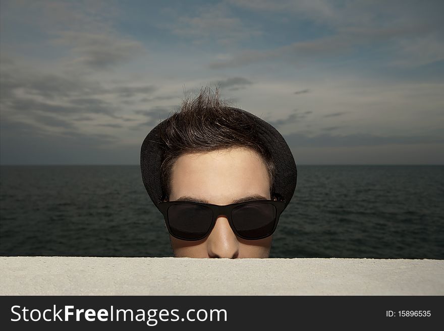 Portrait of weird young man in hat and sunglasses standing near ledge at ocean at sunset