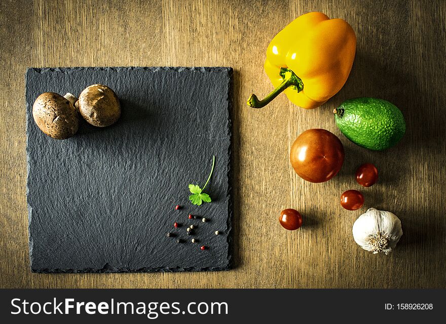 A vegetable composition with pepper, tomato, avocado, garlic and mushrooms on a black chopping board