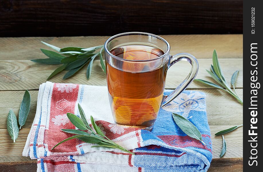 Sage herbal tea or decoction in a glass cup with herb leaves all around on linen blue with red napkin and wooden table, closeup, copy space, alternative medicine and naturopathy concept. Sage herbal tea or decoction in a glass cup with herb leaves all around on linen blue with red napkin and wooden table, closeup, copy space, alternative medicine and naturopathy concept