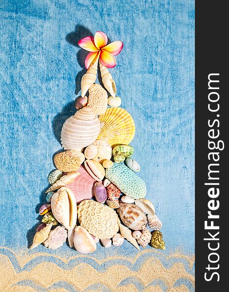 Christmas background with a creative arrangement of seashells, corals and white frangipani flower forming a Xmas tree on blue