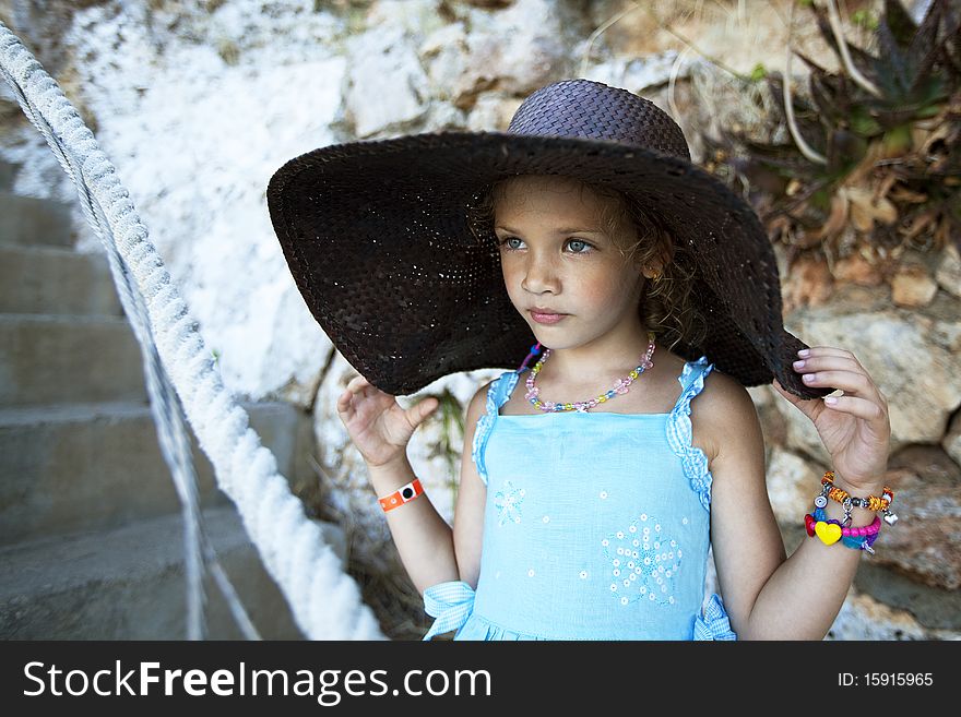 Outdoor portrait of a beautiful little girl with curly hairs in a wide hat on rock background. shot made on a shore of the sea. Outdoor portrait of a beautiful little girl with curly hairs in a wide hat on rock background. shot made on a shore of the sea