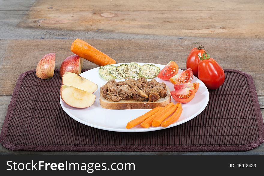 A studio shot of a plated pulled pork sandwich with a quatered apple, carrot, tomatoes and dried cucumber slices as garnishes. A harvest type lunch on a wooden rustic table. A studio shot of a plated pulled pork sandwich with a quatered apple, carrot, tomatoes and dried cucumber slices as garnishes. A harvest type lunch on a wooden rustic table.