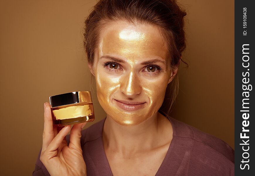 Portrait of smiling middle age woman with golden cosmetic face mask showing cosmetic product jar against bronze background. Portrait of smiling middle age woman with golden cosmetic face mask showing cosmetic product jar against bronze background