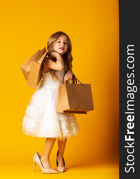 Funny child girl fashionista in big mother`s shoes on yellow background with packages in hand.