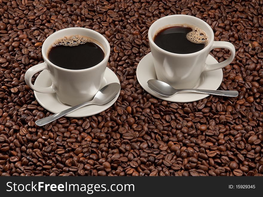 Two cups of black coffee on saucers with coffee spoons standing on roasted coffee beans. Two cups of black coffee on saucers with coffee spoons standing on roasted coffee beans.