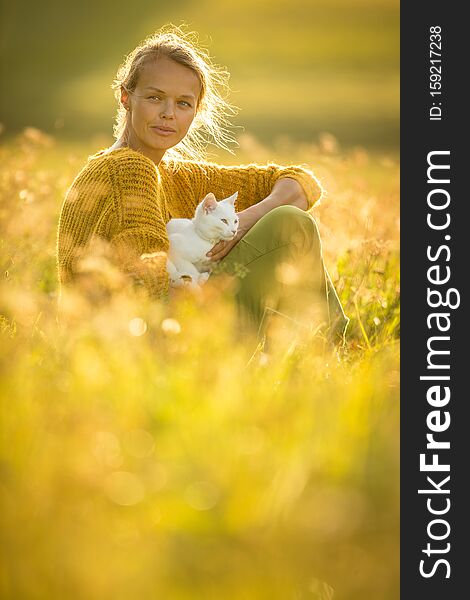 Pretty, young woman with her cat pet sitting in grass on lovely meadow lit by warm evening light shallow DOF; color toned image