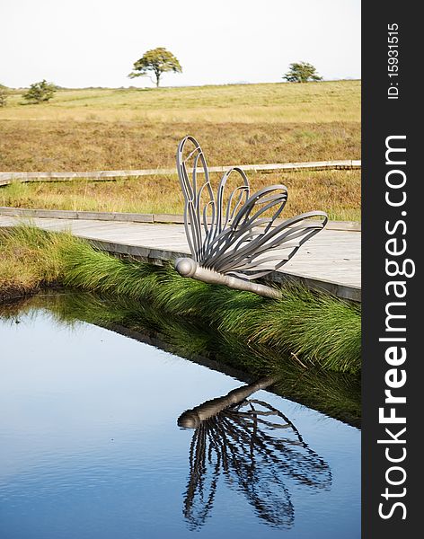 A vertical image of a wooden boardwalk beside a pond with a dragonfly ornament. A vertical image of a wooden boardwalk beside a pond with a dragonfly ornament