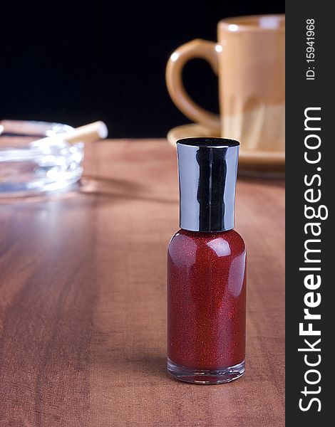 Red nail polish standing on a coffee table in front of a cup of coffee and an ashtray.