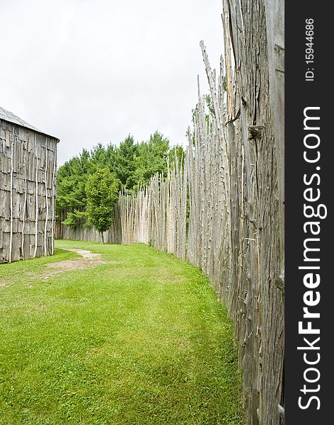 Walkway between a high fence/wall made of bare tree trunks and a long house. Walkway between a high fence/wall made of bare tree trunks and a long house.
