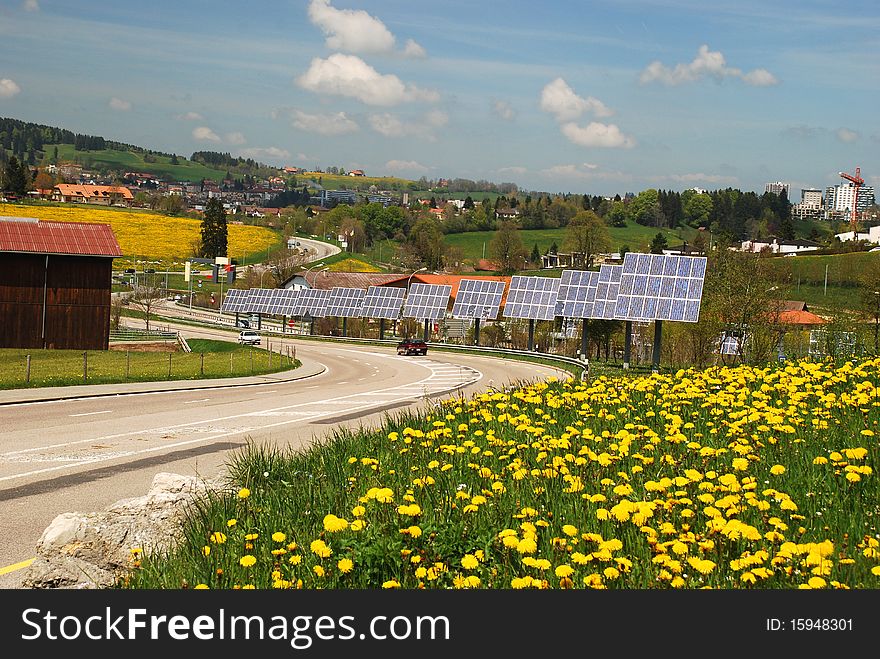 Solar cells along the roat to Le-Chaux-de-Fonds city, yellow flowers of dandelions at foreground. Solar cells along the roat to Le-Chaux-de-Fonds city, yellow flowers of dandelions at foreground