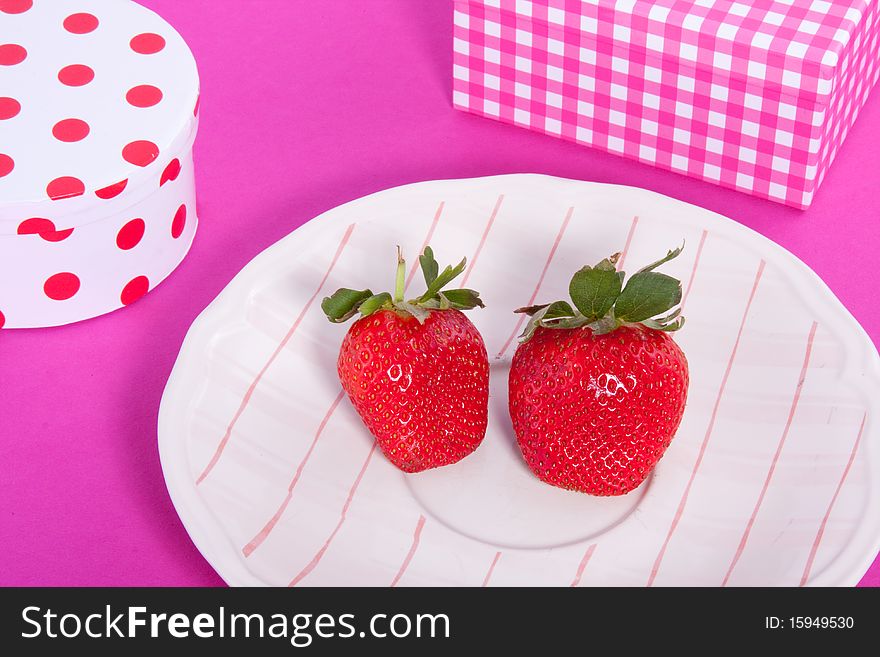 Giftboxes and two red strawberries on a pink white checkered small plate and a pink background. Giftboxes and two red strawberries on a pink white checkered small plate and a pink background