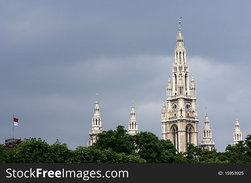 The top part of towers of a city town hall in Vienna and the Austrian flag over crones of trees against the storm sky. The top part of towers of a city town hall in Vienna and the Austrian flag over crones of trees against the storm sky