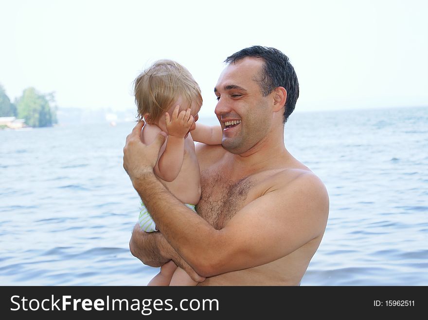 In the summer on a beach the laughing father holds on hands of the small child and plays with it. In the summer on a beach the laughing father holds on hands of the small child and plays with it