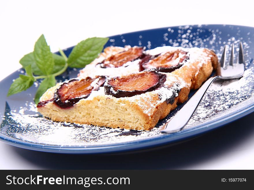 Piece of Plum Pie covered with powder sugar on a blue plate decorated with a mint twig