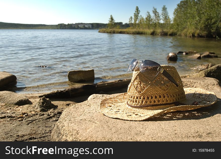 The straw hat and sun glasses lie on a stone on the bank of lake