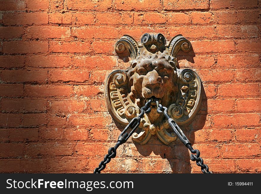 A rusty, lion faced gargoyle on a brick wall with a chain in it's mouth. The gargoyle is illuminated by a ray of light. A rusty, lion faced gargoyle on a brick wall with a chain in it's mouth. The gargoyle is illuminated by a ray of light.