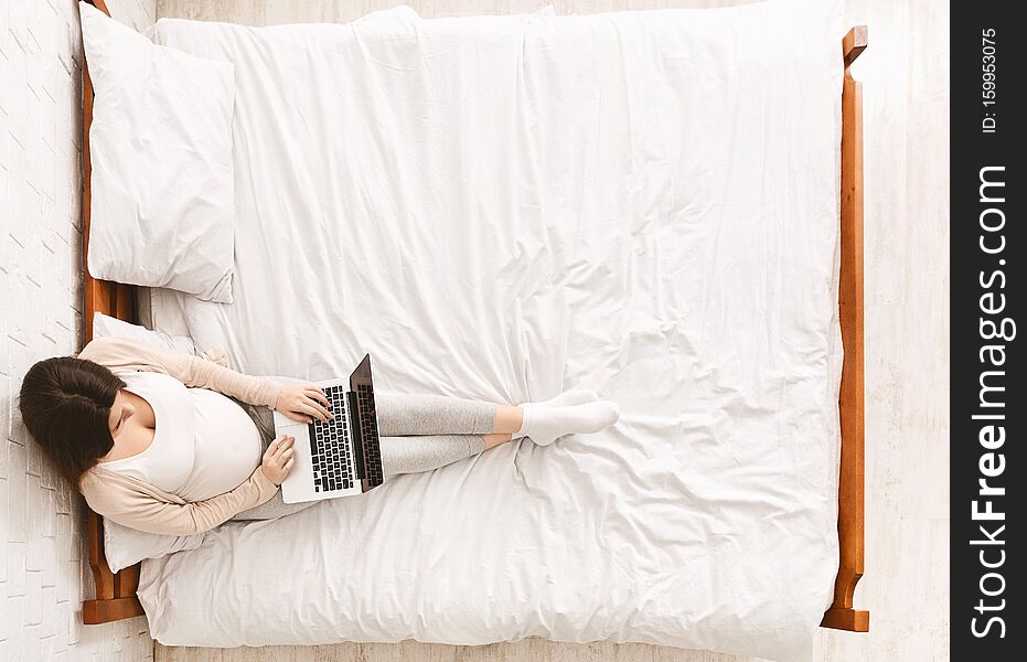 Pregnant woman working on laptop, sitting on bed