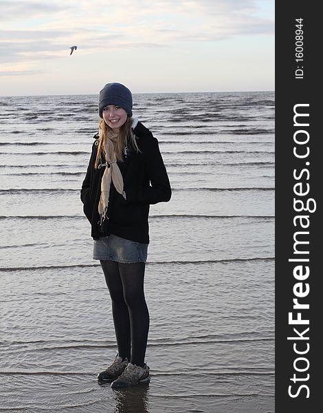 Girl in hat and scarf standing at the waters edge on a beach in winter with seagull in background. Girl in hat and scarf standing at the waters edge on a beach in winter with seagull in background