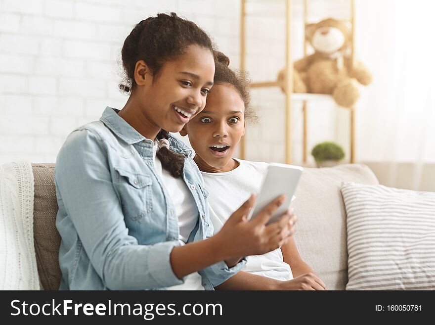 Shocking mobile app. African girl showing her little sister new mobile application, sitting at home, copy space. Shocking mobile app. African girl showing her little sister new mobile application, sitting at home, copy space