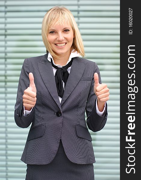 A young and attractive blond businesswoman in a formal dress is holding thumbs up as a sign of success. The image is taken on an abstract background.