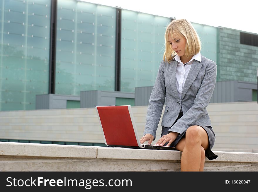 A young and attractive business woman in formal clothes is working outdoors with a laptop. The image is taken on a modern background. A young and attractive business woman in formal clothes is working outdoors with a laptop. The image is taken on a modern background.