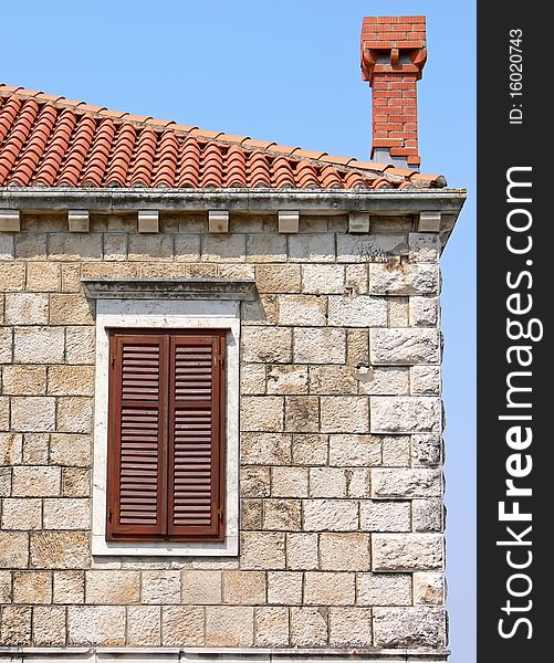 Part of the old stone building. Closeup of the window with shutters on the facade, red tiled roof and bricked chimney. Part of the old stone building. Closeup of the window with shutters on the facade, red tiled roof and bricked chimney.