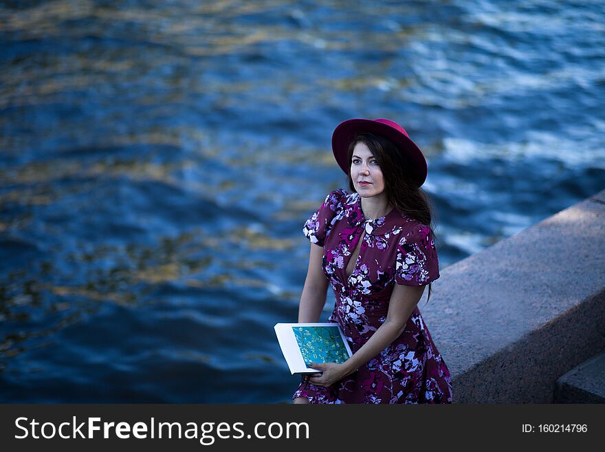 A Young Woman In A Dress And Hat, Holding A Notebook