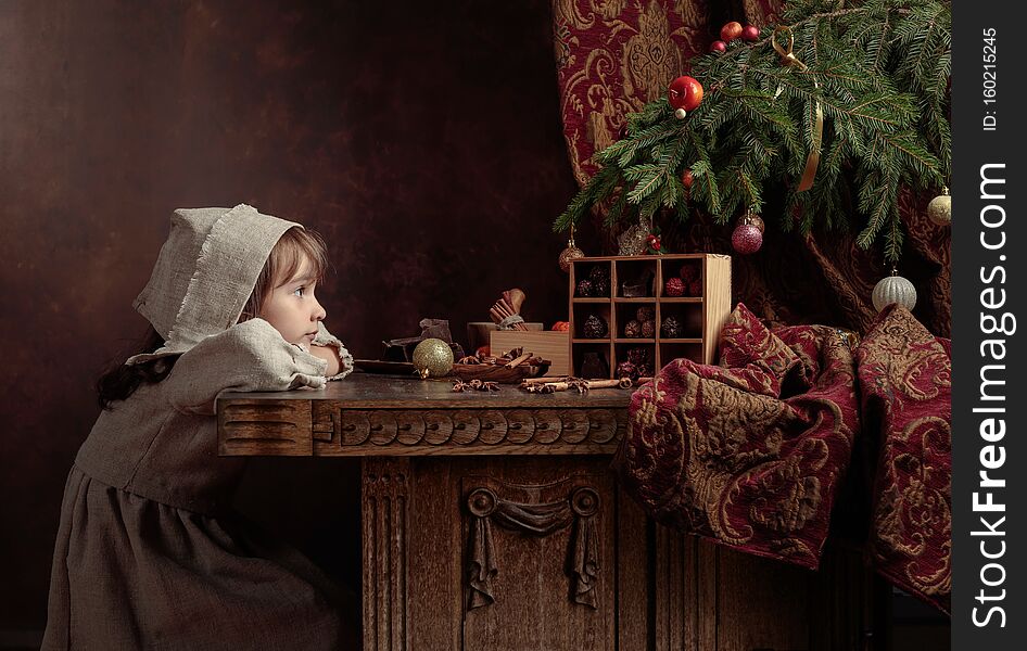 Little girl in an vintage linen dress dreaming near the table with sweets. Chocolates and spices on the table under the Christmas tree. Genre portrait in retro style. Little girl in an vintage linen dress dreaming near the table with sweets. Chocolates and spices on the table under the Christmas tree. Genre portrait in retro style