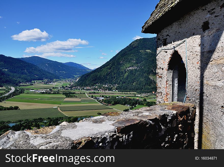 Image shows a panoramic view to Carinthia from Castle Landskron, Austria. In front there are some details of Castle Landskron, meadows and fields in the middle, in the background there are some mountains and cloudy blue sky. Image shows a panoramic view to Carinthia from Castle Landskron, Austria. In front there are some details of Castle Landskron, meadows and fields in the middle, in the background there are some mountains and cloudy blue sky.