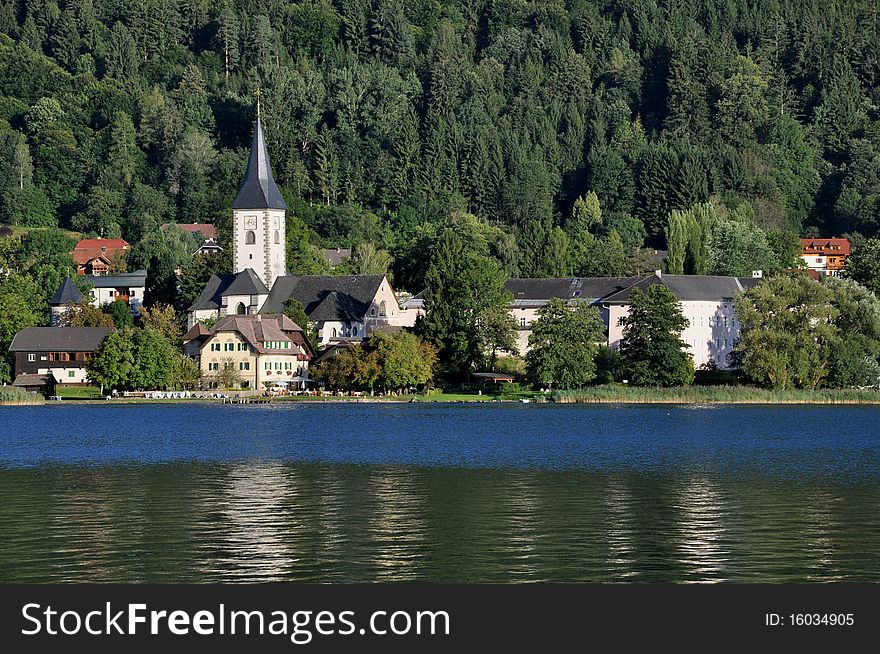 Image shows the Ossiach Abbey, Austria in front of green forest. In the front lake Ossiach, Austria. Ossiach Abbey is a former Benedictine monastery.