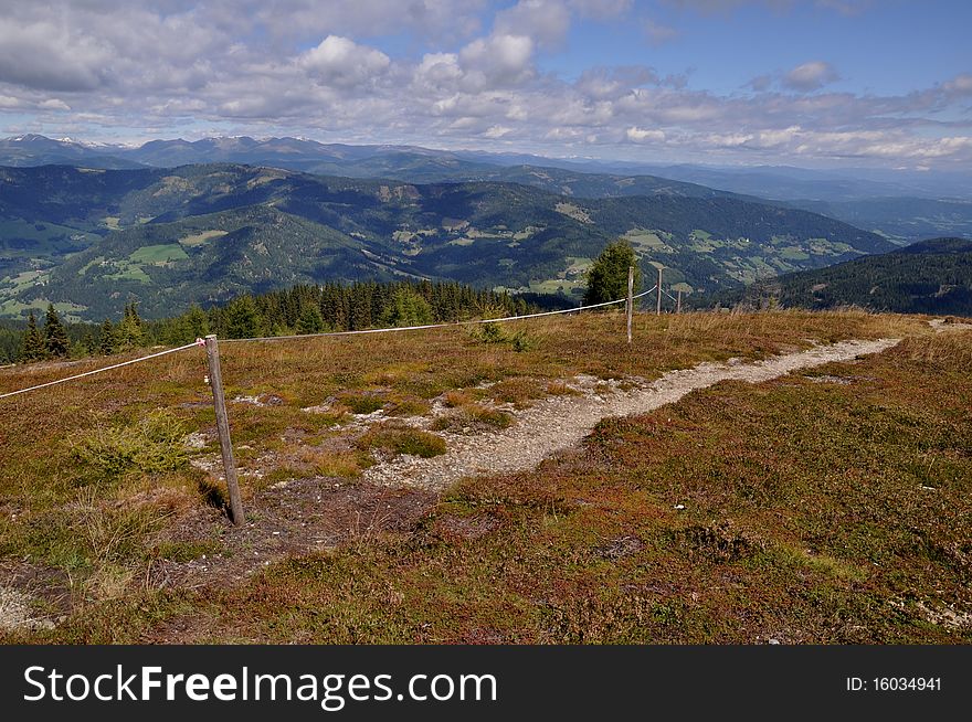 Panoramic view from mountain Gerlitzen to Carinthia, Austria. Image shows a hiking trail with meadow and a fence. In the background is the landscape of Carinthia, Austria. Sky is blue and cloudy. Panoramic view from mountain Gerlitzen to Carinthia, Austria. Image shows a hiking trail with meadow and a fence. In the background is the landscape of Carinthia, Austria. Sky is blue and cloudy.