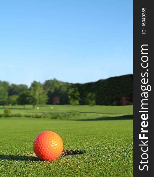 Orange gold ball on the green beside the hole with clear blue sky in background.