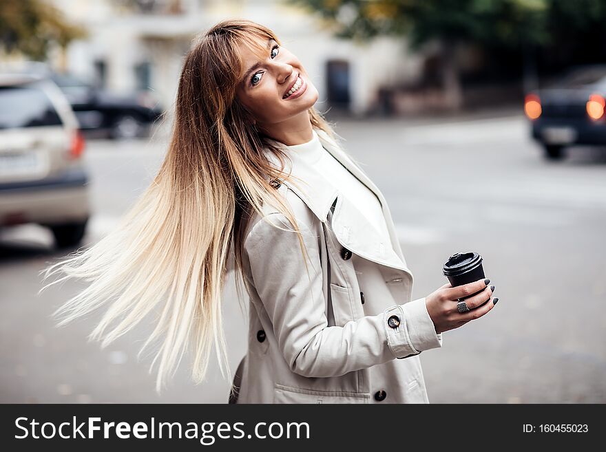 Stylish beautiful woman blonde in beige trench coat holds coffee to go. portrait of smiling girl . Street fashion concept.Dynamically young girl walks down the street.Hair fluttering in the wind. Stylish beautiful woman blonde in beige trench coat holds coffee to go. portrait of smiling girl . Street fashion concept.Dynamically young girl walks down the street.Hair fluttering in the wind