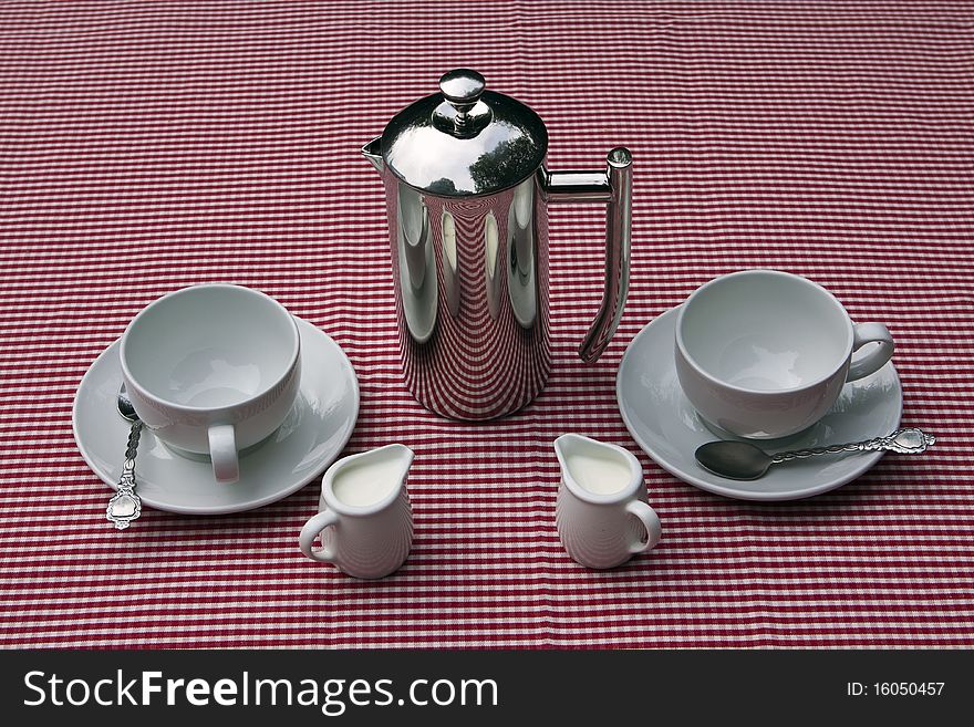 A shiny coffee pot and cups and saucers on a red and white tablecloth