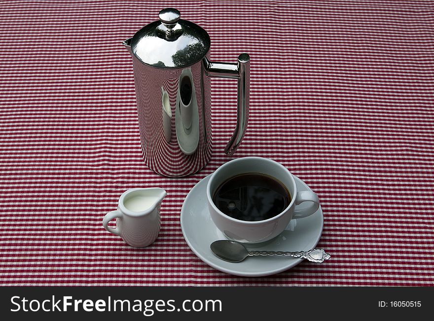 A shiny coffee pot and cups and saucers on a red and white tablecloth