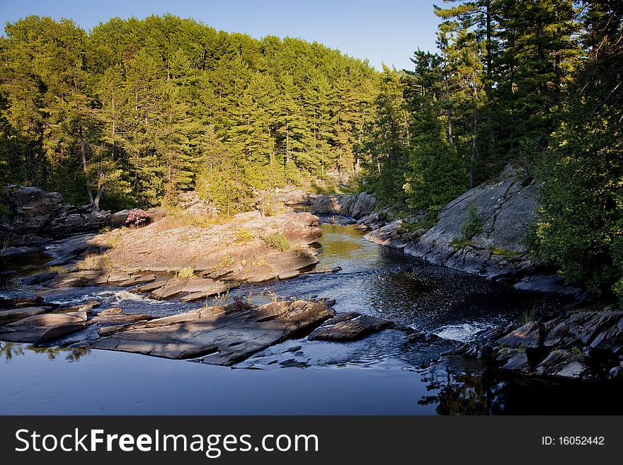 Shot from above falls, looking down river towards a sunlit forest. The river itself is mostly in shadow. Shot in Northern Ontario, Canada. Shot from above falls, looking down river towards a sunlit forest. The river itself is mostly in shadow. Shot in Northern Ontario, Canada.