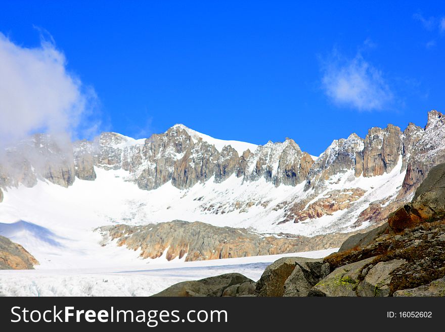 A snow capped mountain in Switzerland and a rocky hillside with clouds coming in. A snow capped mountain in Switzerland and a rocky hillside with clouds coming in
