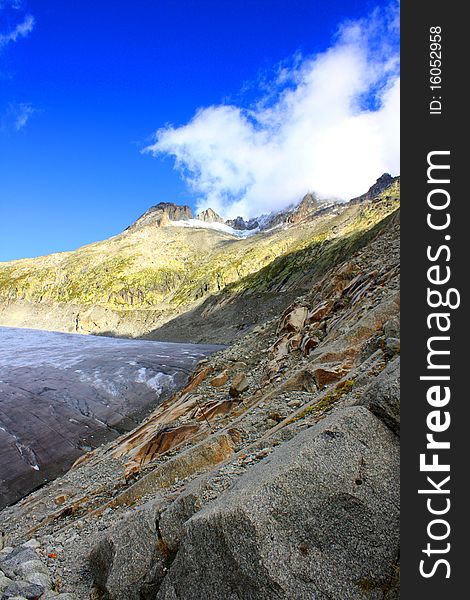 A rocky mountain side with snow cappe mountains and a glacier with clouds and a bright blue sunny sky. A rocky mountain side with snow cappe mountains and a glacier with clouds and a bright blue sunny sky