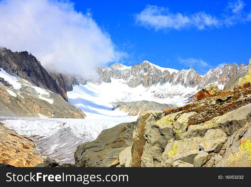 Rocky mountain side covering a glacier with snowy mountains in the back under a blue sunny sky and clouds creeping in. Rocky mountain side covering a glacier with snowy mountains in the back under a blue sunny sky and clouds creeping in