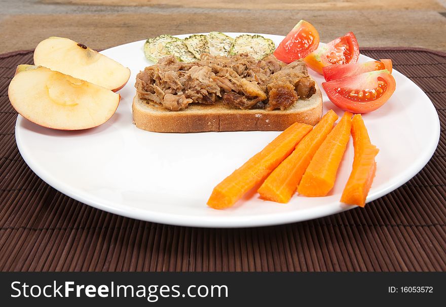 A wide angle studio shot of a plated pulled pork sandwich with side dishes of carrots,tomatoes,apples and dried cucumber slices. A wide angle studio shot of a plated pulled pork sandwich with side dishes of carrots,tomatoes,apples and dried cucumber slices.