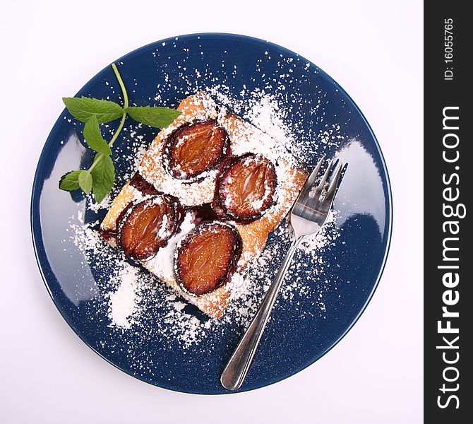 Piece of Plum Pie covered with powder sugar on a blue plate decorated with a mint twig